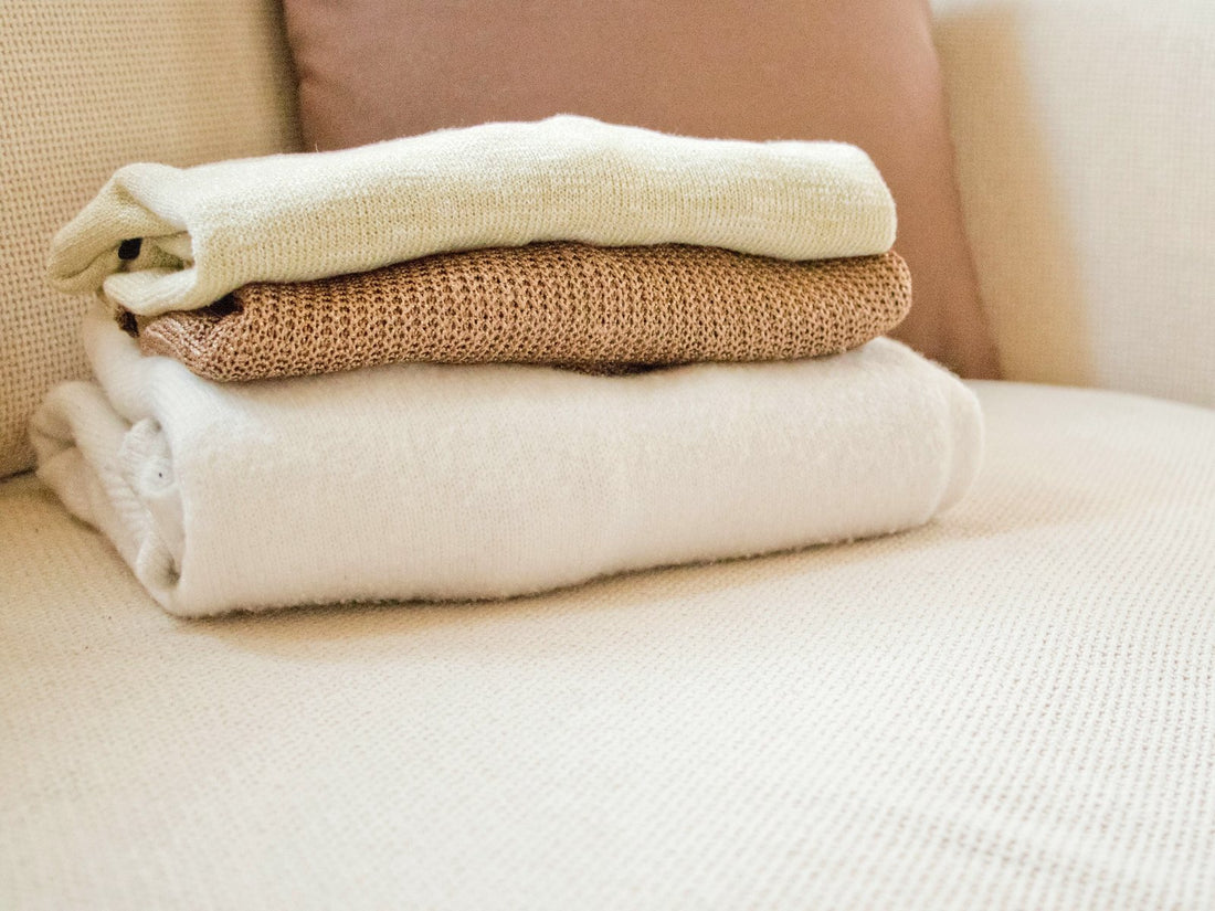Three folded knit sweaters in ecru, camel and white on a cream sofa